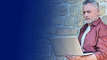 Older man using laptop near a stone wall outdoors.